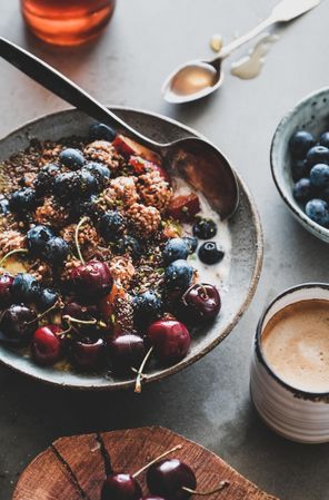 Oat granola yogurt bowl with cherries, blueberries, nuts, coffee, wooden board in foreground