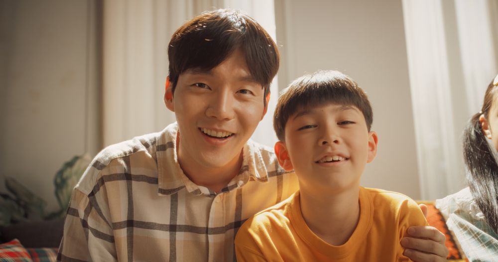 Portrait of a Happy Korean Father and Son in the Living Room at Home, Smiling and Waving at the Camera.