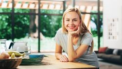 Portrait of Young Adult Woman with Blond Hair Wearing Gray V-Neck T-Shirt, Leaning on a Table and Smiling Charmingly
