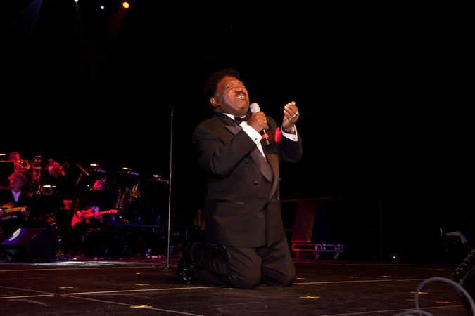 Black male artist Percy Sledge singing and kneeling on stage with band