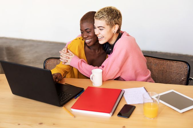 Female friends embracing happily over laptop at breakfast table