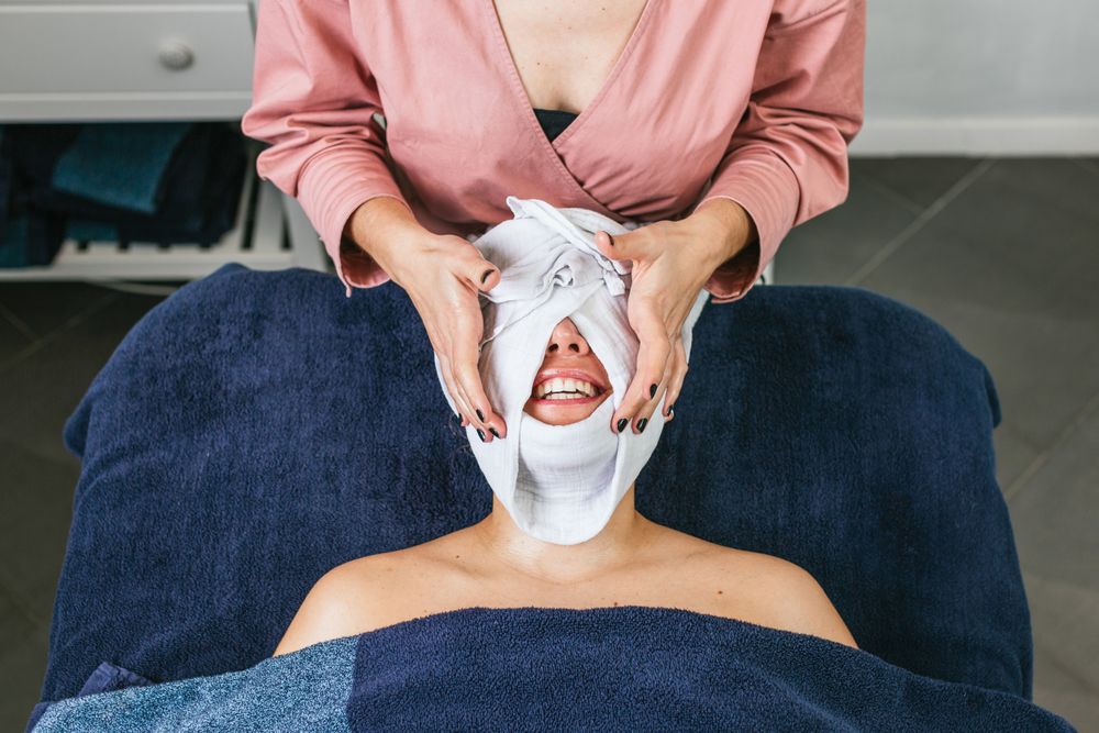Crop cosmetologist steaming face of client with hot towel