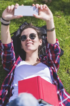Above view of a young smiling hipster woman lying on grass in a sunny day at a park while taking a selfie with a mobile phone