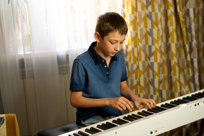Boy in blue shirt playing digital piano near window