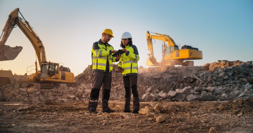 White Male Civil Engineer Talking To Hispanic Female Inspector And Using Tablet On Construction Site of New Apartment Complex. Real Estate Developers Discussing Business, Excavators Working.