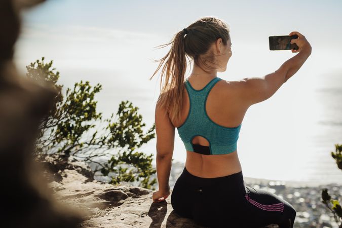 Rear view of a fit young woman sitting on a cliff taking selfie