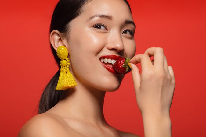 Female model with long earring and eating strawberry against  red background