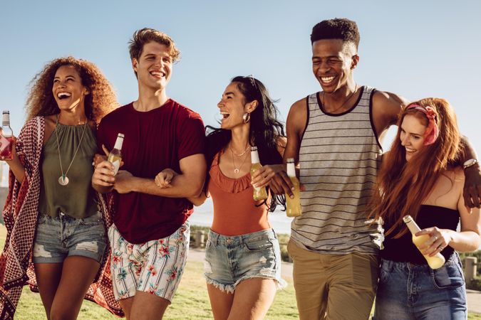 Group of friends walking together with locked arms while holding beers in hand