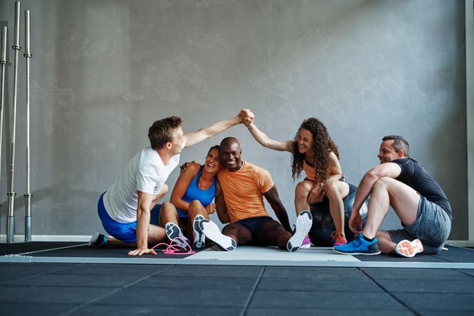 Multi-ethnic group of people in workout gear sitting on gym floor with two people high fiving