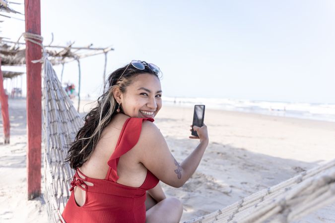 A young Hispanic woman is looking at the camera while taking a photo of the sea on a hammock at the beach