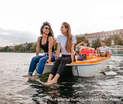 Two women friends sitting in front pedal boat with feet in water and man in background bGozlb