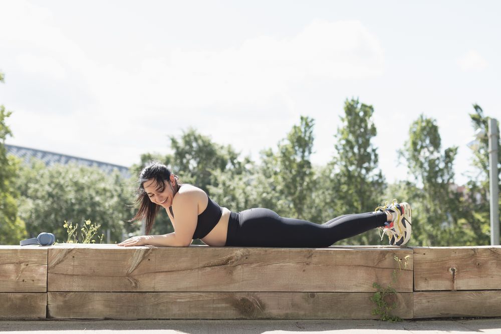A young woman laughs happily as she relaxes between workouts, lying on a bench in the park