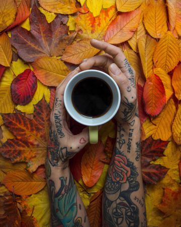 Person with tattooed arms holding a cup of coffee over yellow tree leaves