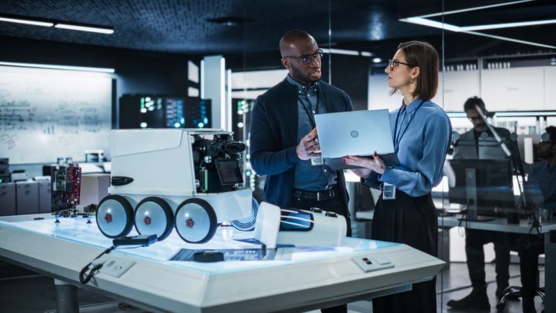 Two Diverse Engineers Developing a Remotely Controlled Mobility Robot in an Industrial Research Facility. Black Male Specialist Pitching his Ideas and Advising his Colleague Using Laptop Computer
