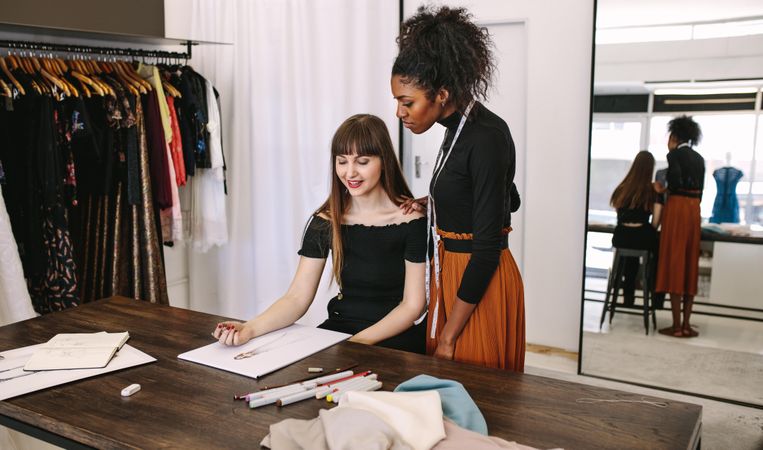 Fashion designer sketching a design sitting at her table while another woman looks on