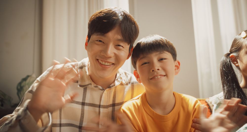 Portrait of a Happy Korean Father and Son in the Living Room at Home, Smiling and Waving at the Camera.