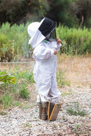 Child in protective costume standing near bee smoker