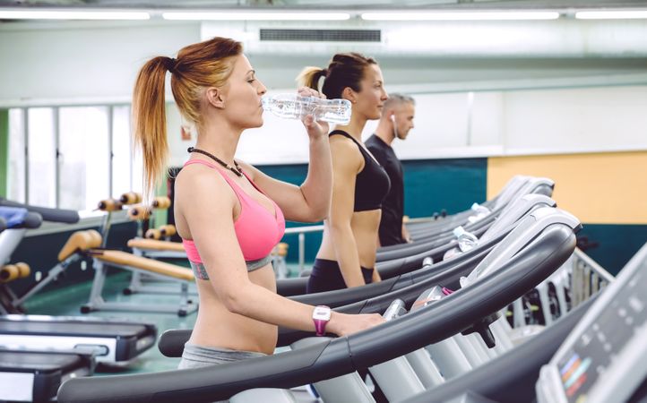 Side view of woman drinking water while working out on treadmill in gym