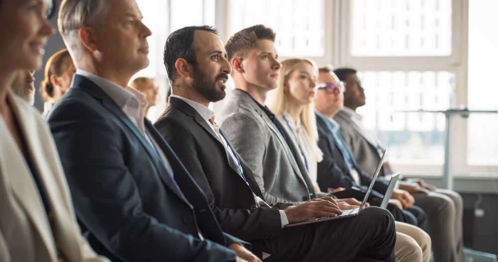 Multiethnic Man Sitting in Crowded Auditorium at Conference. Middle Eastern Businessman Listening To Keynote And Typing On Laptop. Specialist Watching Technology Presentation About Software Solutions.