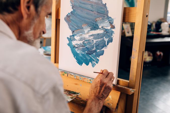 Older male artist working on corner of easel of his painting