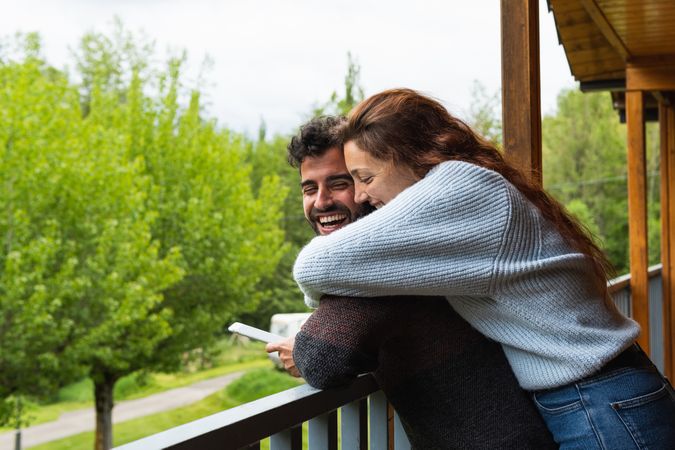 Cheerful traveling couple embracing and using smartphone on terrace of house