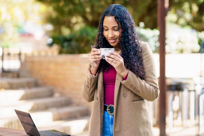 A woman is enjoying her coffee while diligently working on her laptop outdoors amidst nature