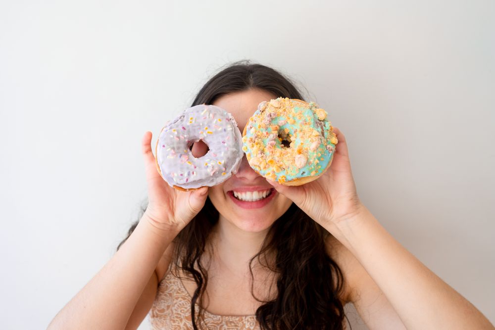 Playful woman making glasses of donuts while standing by white wall