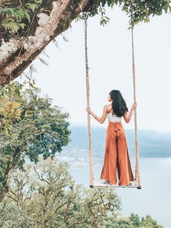 Back view of young woman standing on swing chair hanging from tree in Bali, Indonesia