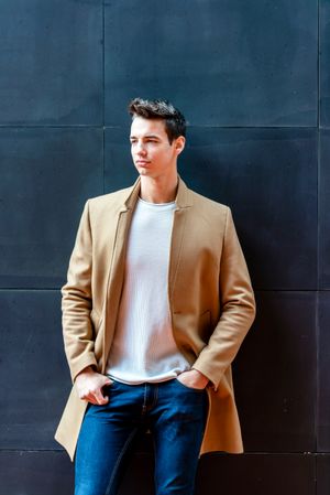 Stylish man leaning on dark wall outside looking away from camera dressed in camel coat 