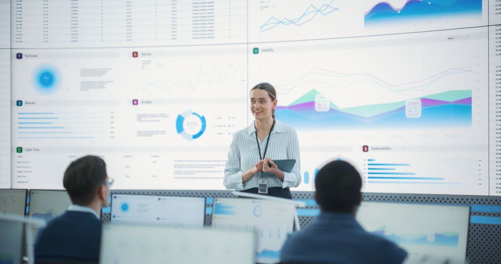 Female Project Manager With Tablet Computer Is Giving Presentation To Diverse Employees In Front of Big Digital Screen In Monitoring Room.