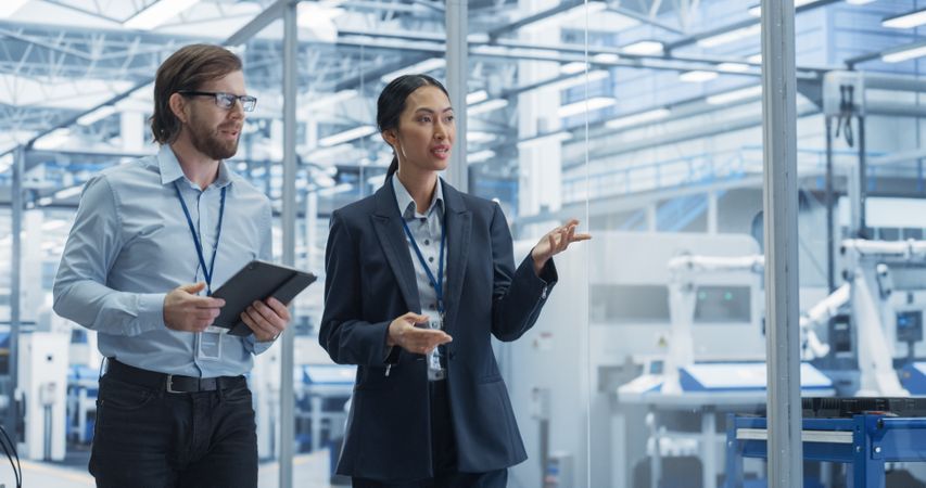 Two Diverse Engineers Walking in Office, Using Tablet Computer at a Modern Electronics Factory. Female and Male Specialists Chatting, Monitoring Robots on Modern Assembly Line.