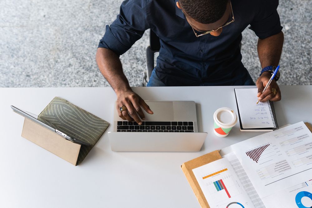 Black male writing in notepad near laptop in office