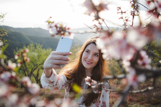 Young happy woman taking selfie on smartphone near trees on hill