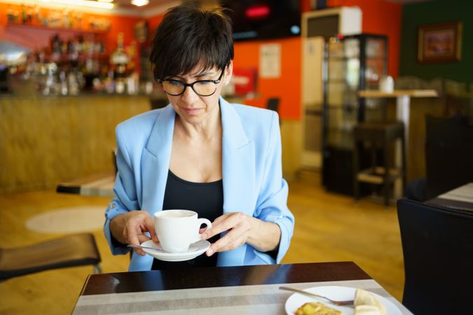 Woman in glasses and blue blazer looking down at her coffee in cafe