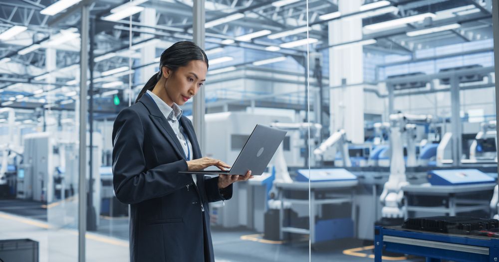Asian Female Engineer Standing Next to a Window in Office, Using Laptop Computer at Electronics Factory. Machines are Undergoing Maintenance, Specialist Using an Online Software