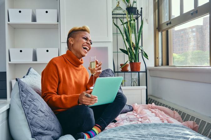 Joyful Black woman laughing at a laptop with champagne flute in a bright modern apartment