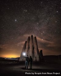Silhouette of two people holding a torch standing beside hand shaped rock formation under starry night 0WJD6b