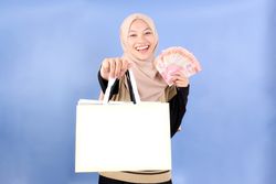 Cheerful Asian Muslim Woman in Hijab Holding Blank Shopping Bag for Mock Up with Rupiah Cash