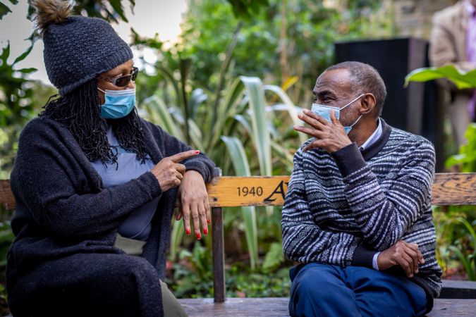 Mature Black man and woman sitting outside talking in facemasks