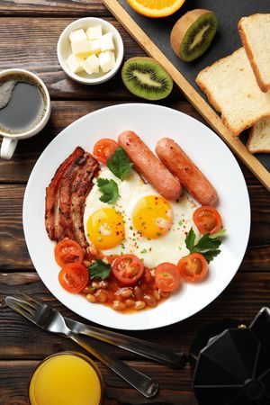 Wooden table with breakfast plate of coffee eggs, tomatoes, sausage and bacon, vertical