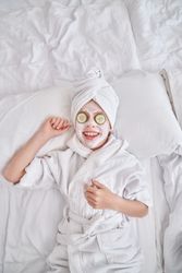 Kid in bathrobe and facial mask relaxing on bed at home