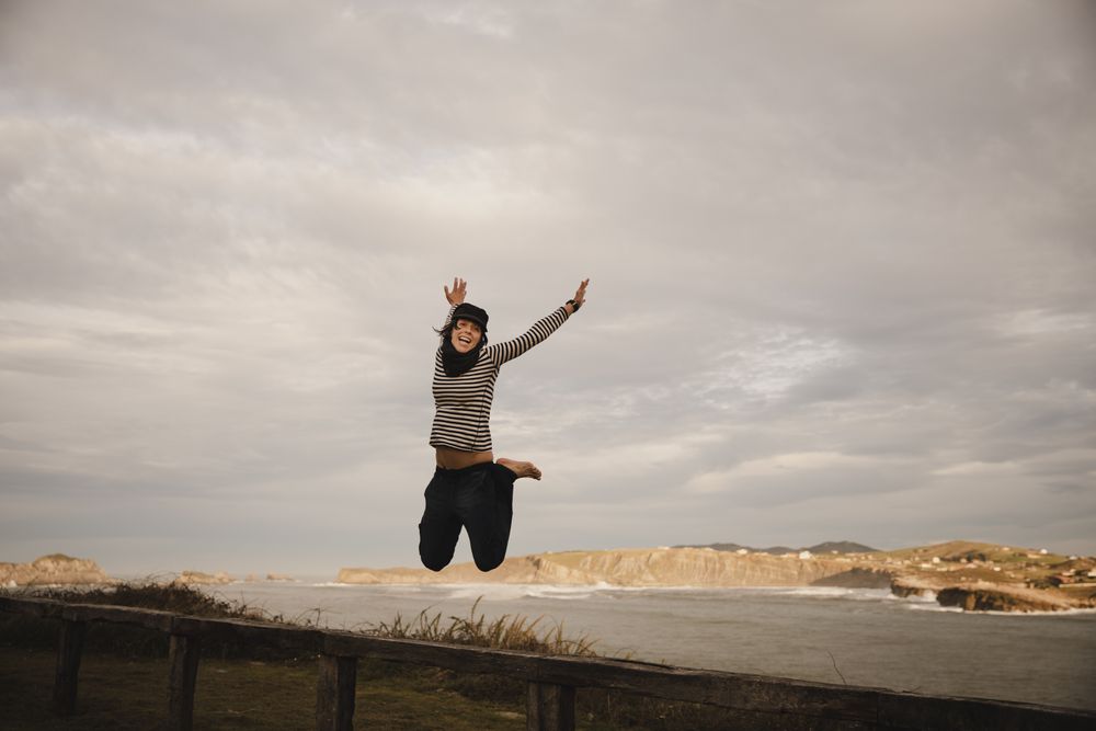 Young woman in hats having fun on seat on coast near waving sea and cloudy sky