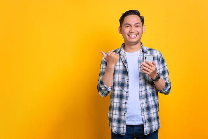 Smiling Asian male pointing thumb to the side while holding smart phone
