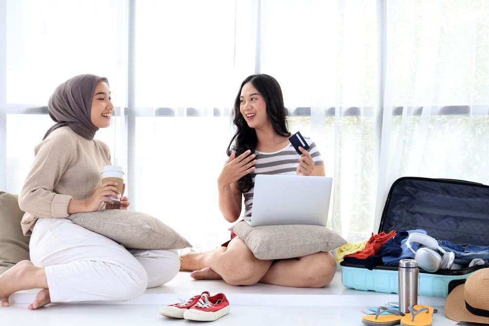Two Asian Friends Discuss Vacation Ticket Payments While Packing Their Suitcases