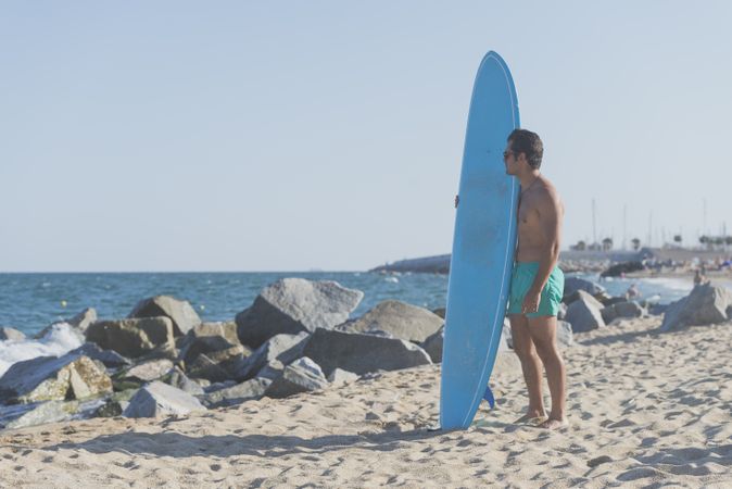 Full body of male surfer holding surfboard standing and looking out at ocean