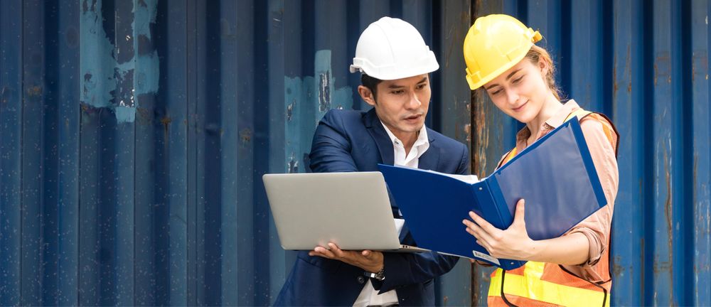 Two colleagues in hard hats discussing logistics on dock near containers