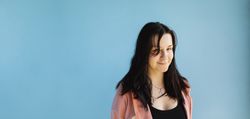 A young person with long hair, on a plain blue background, looks at the camera confidently