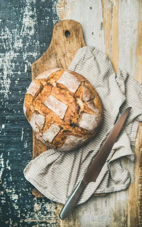 Scored homemade loaf of bread on bread board with linen napkin and serrated knife