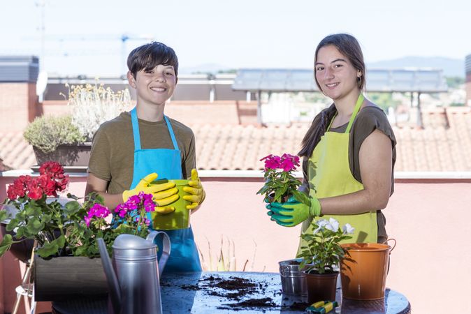 Two happy teenager wearing gardener apron posing at home terrace while holding a plant pot