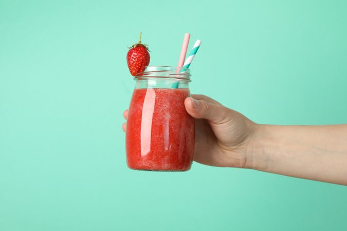 Female hand holds glass jar with strawberry cocktail on mint background. Summer drink